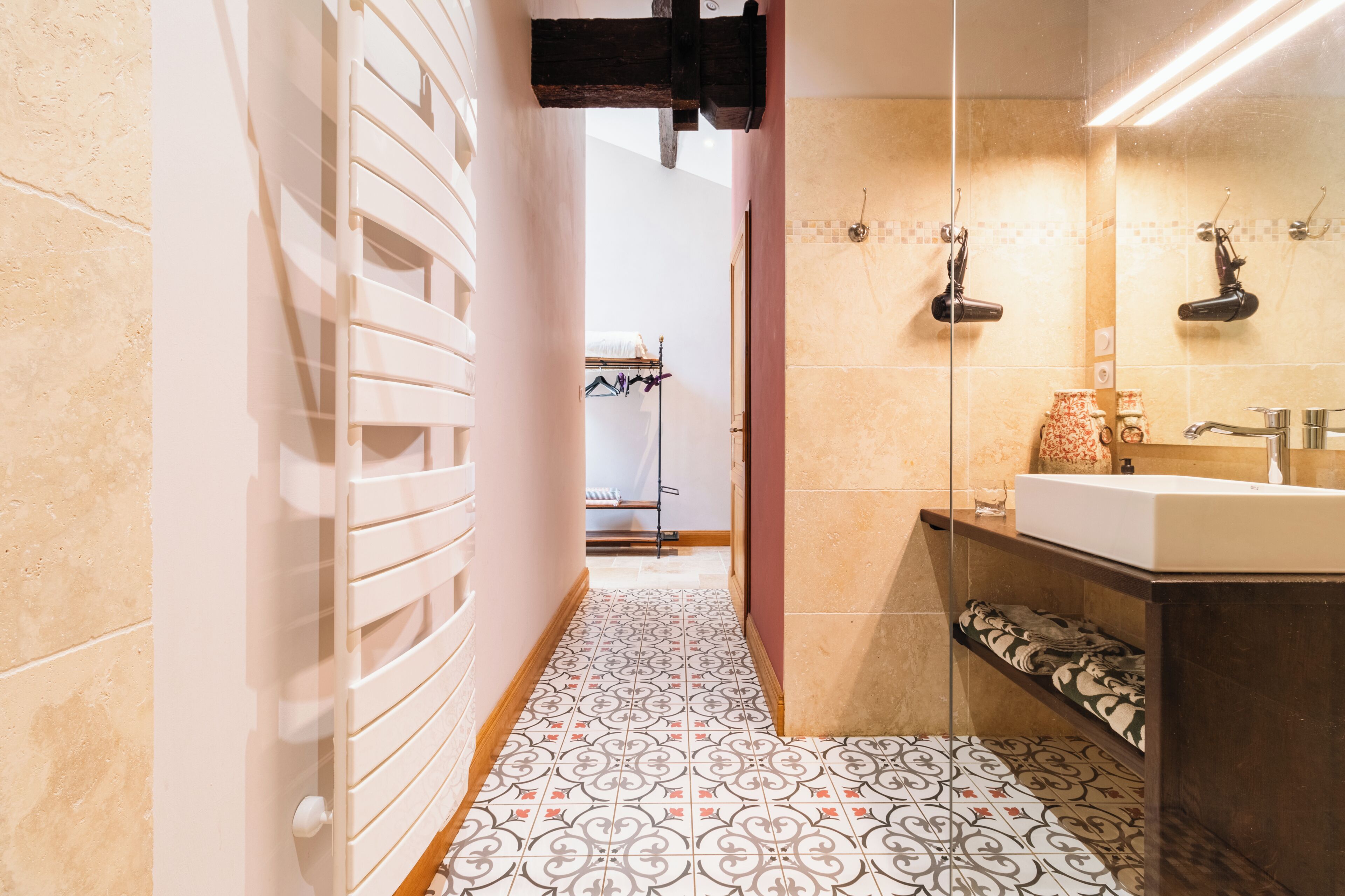 Un couloir étroit avec des carreaux de sol à motifs, un radiateur blanc et une salle de bain équipée d’un lavabo et d’un miroir.