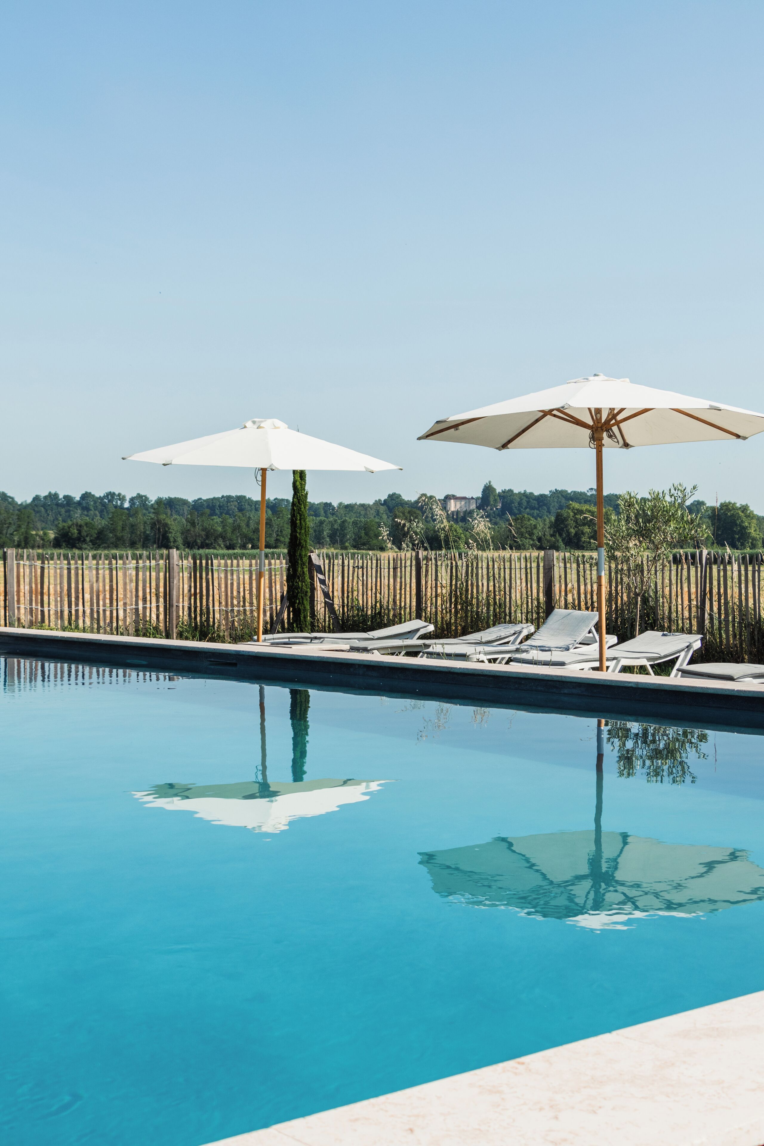 A swimming pool with lounge chairs and umbrellas, surrounded by a wooden fence and trees.