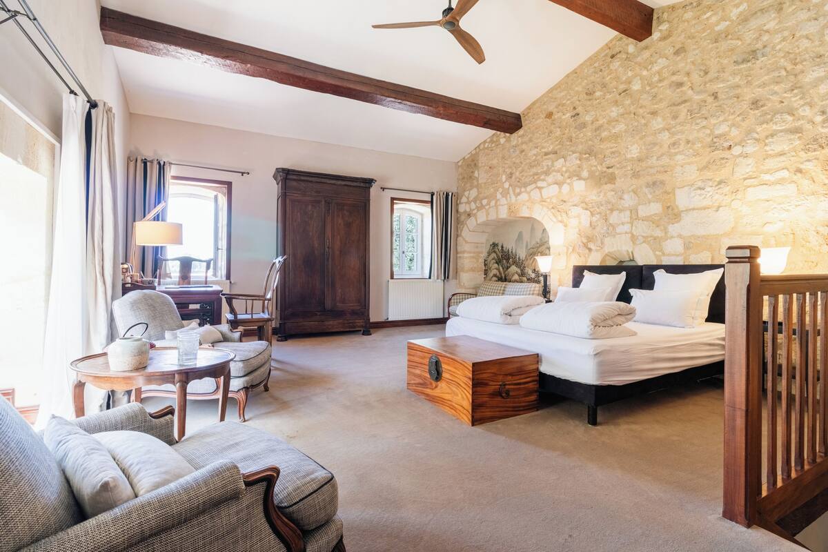 A room with a bed, a wooden chest, a sofa, a chair, and a stone wall.