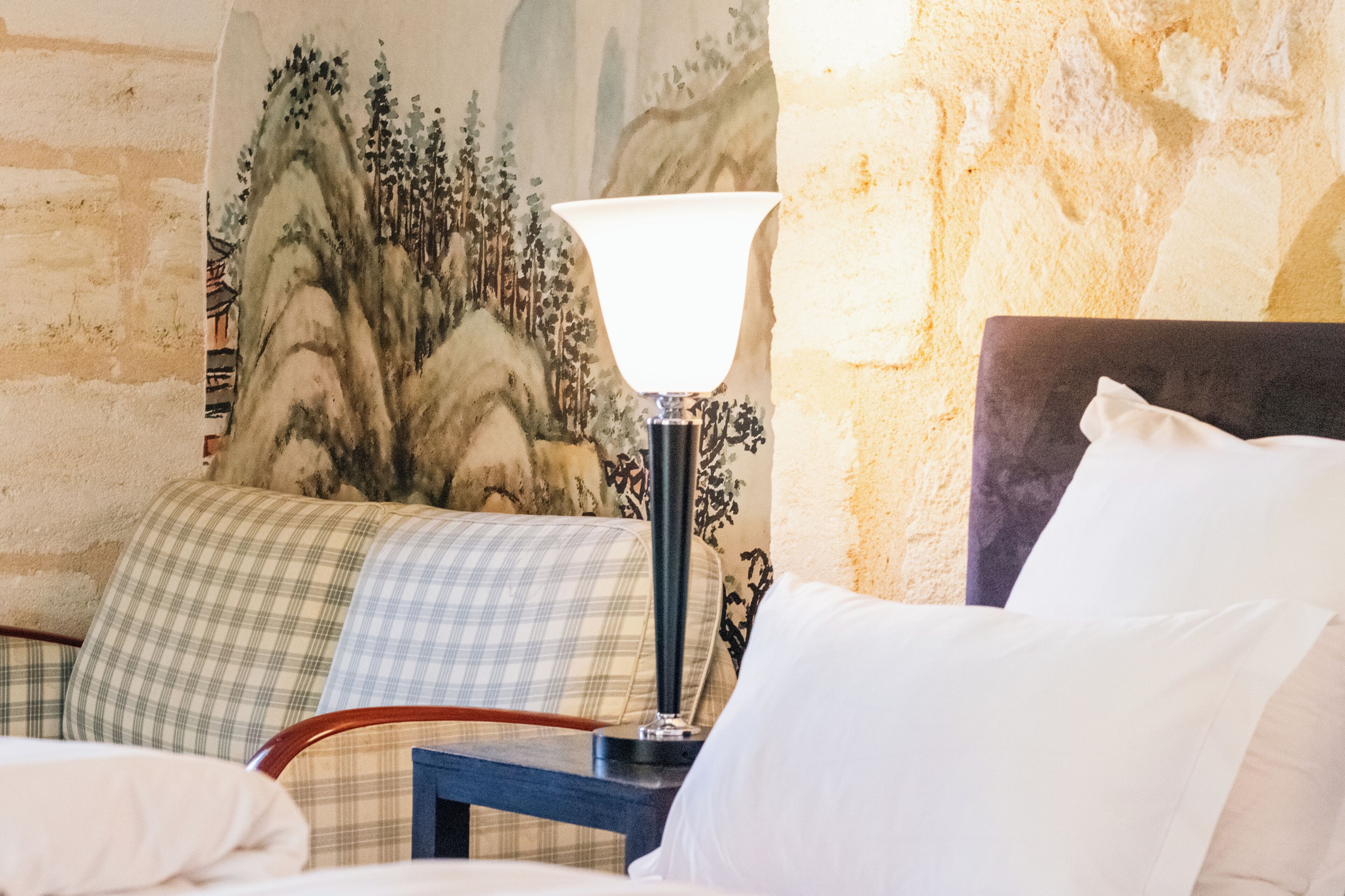 A room with a bed, a nightstand with a lamp, a checkered blanket, and a wall with a painted landscape.