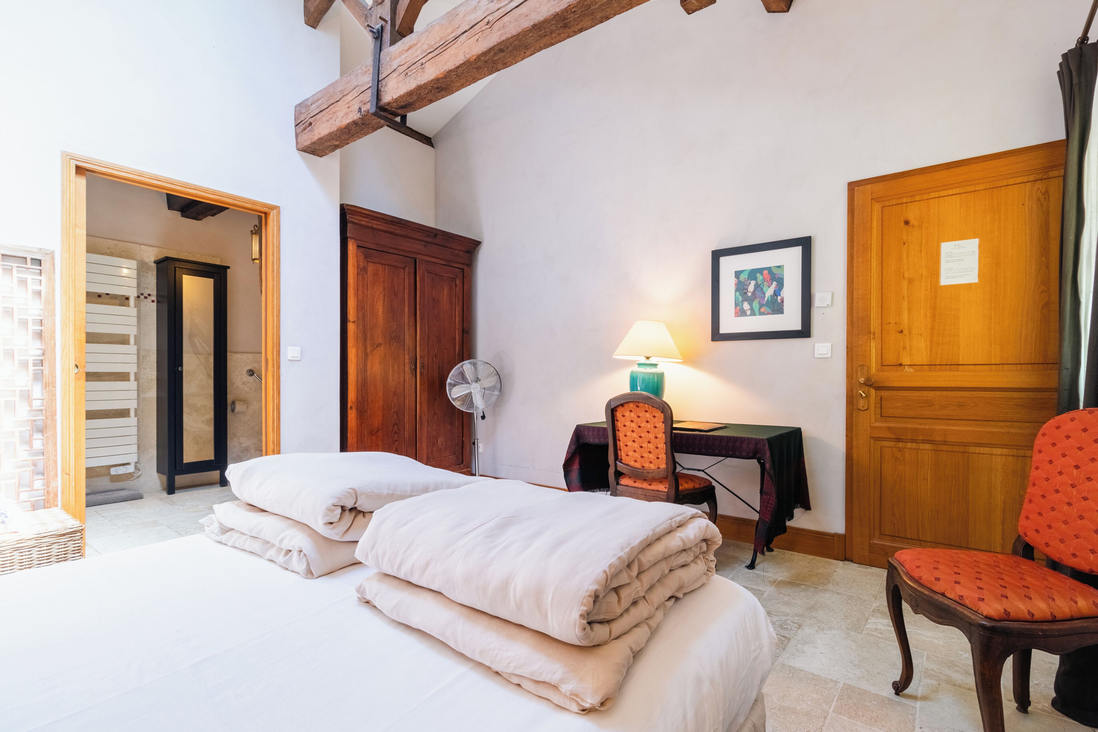 Un dormitorio con cama, almohadas, un escritorio con lámpara, una silla y una puerta de madera.