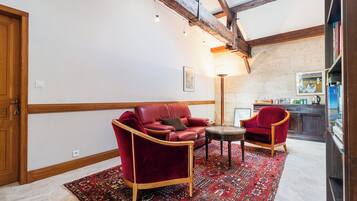 Een kamer met een rode leren bank, een bijpassende fauteuil, een klein rond tafeltje en een kamerbreed tapijt met dessin. Er is een houten balk in het zicht, een wandlamp en een keuken in de achtergrond.
