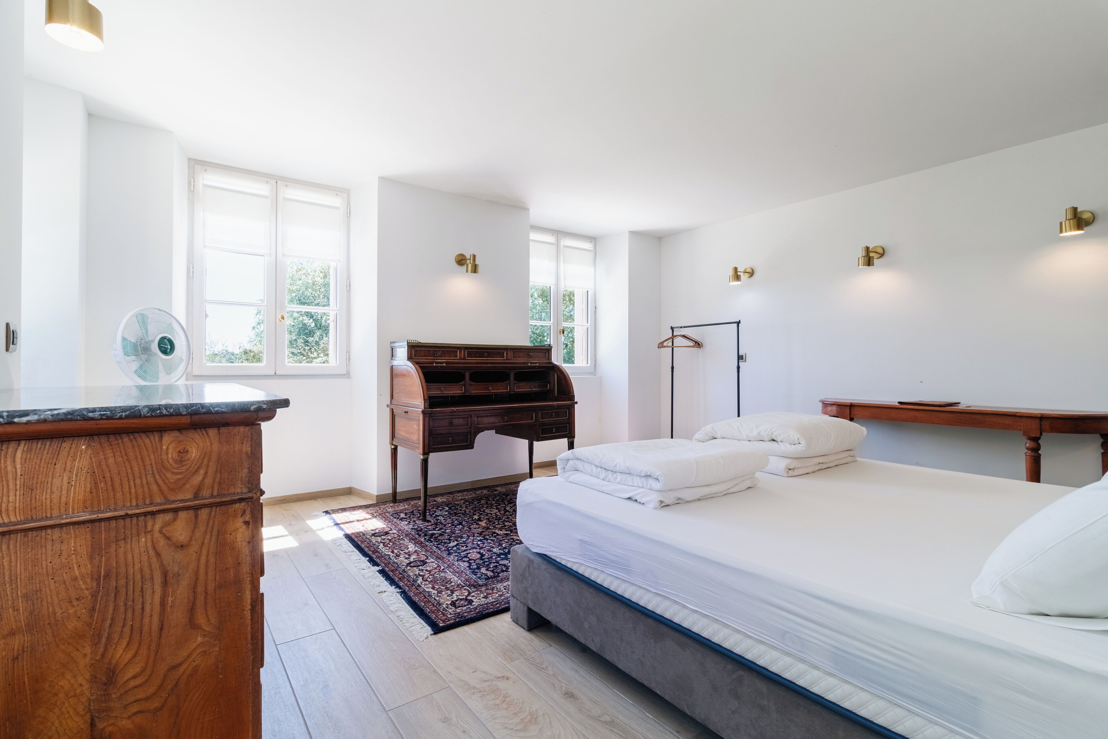 A bedroom with a bed, dresser, desk, and a standing fan.