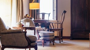 Una habitación con un ventanal grande, una lámpara, una silla de madera, una mesa y una estantería.