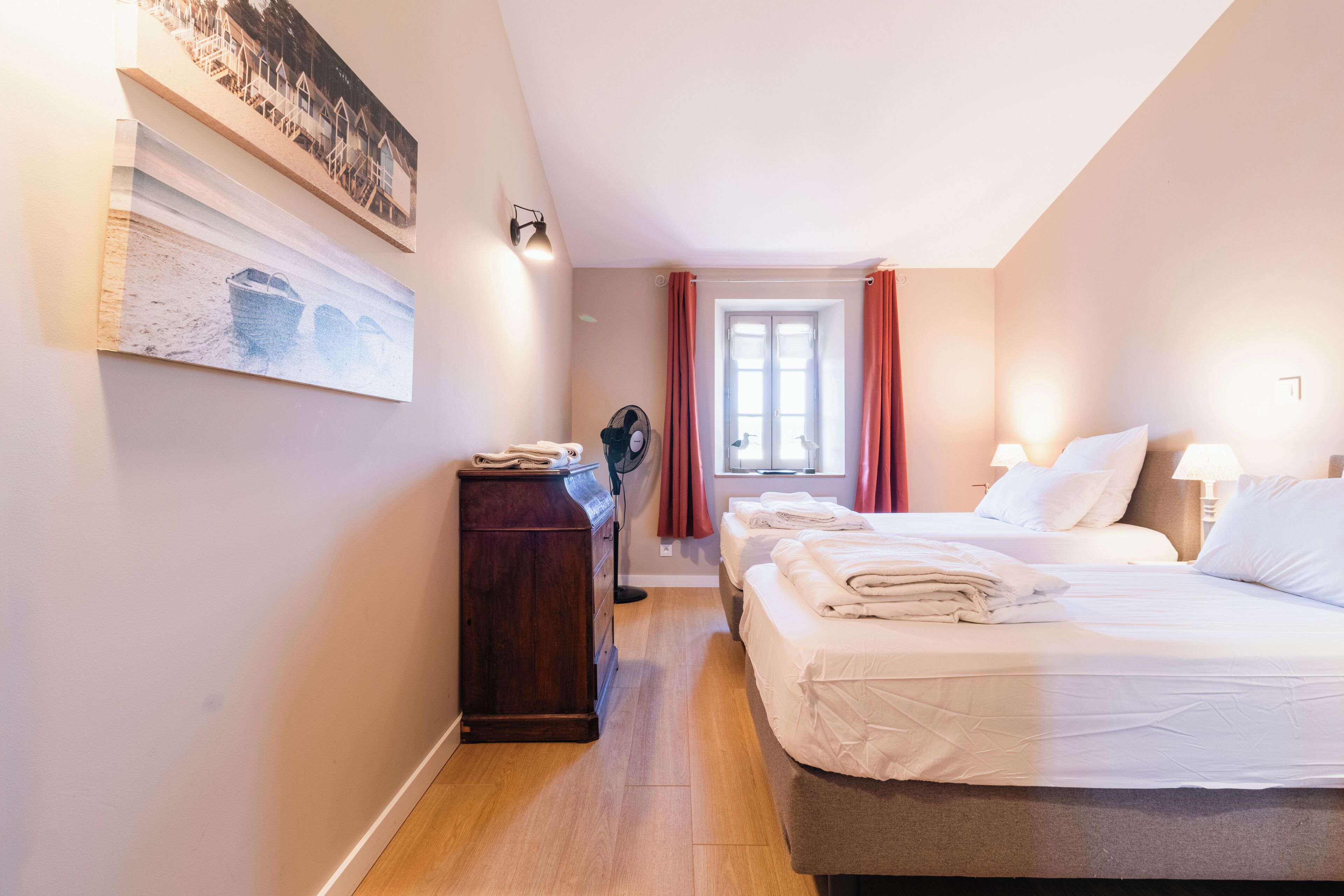 Quarto de hotel com cama, penteadeira, ventilador, janela com cortinas e obras de arte na parede.