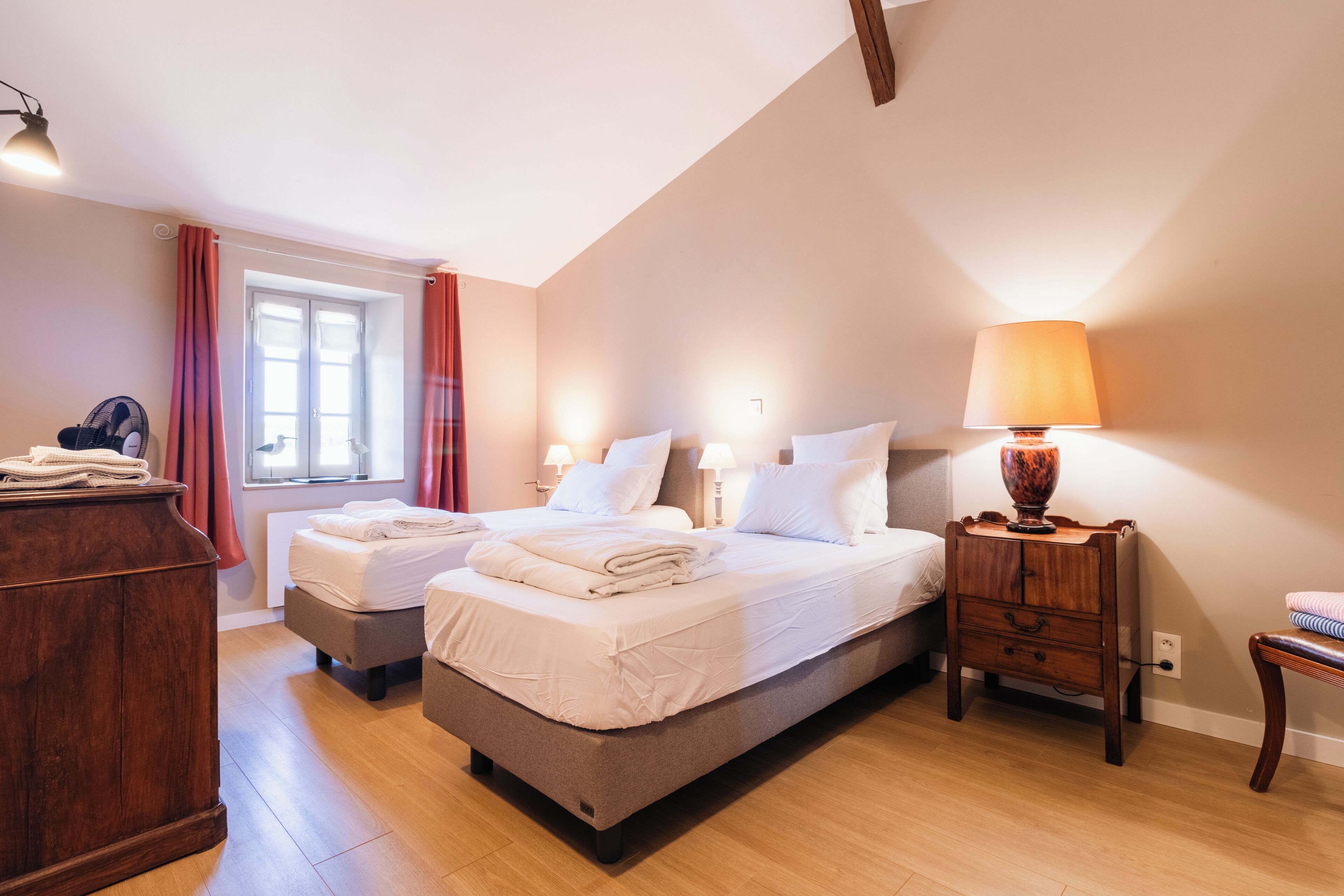 A bedroom with two beds, a wooden dresser, a bedside table with a lamp, and a window with curtains.