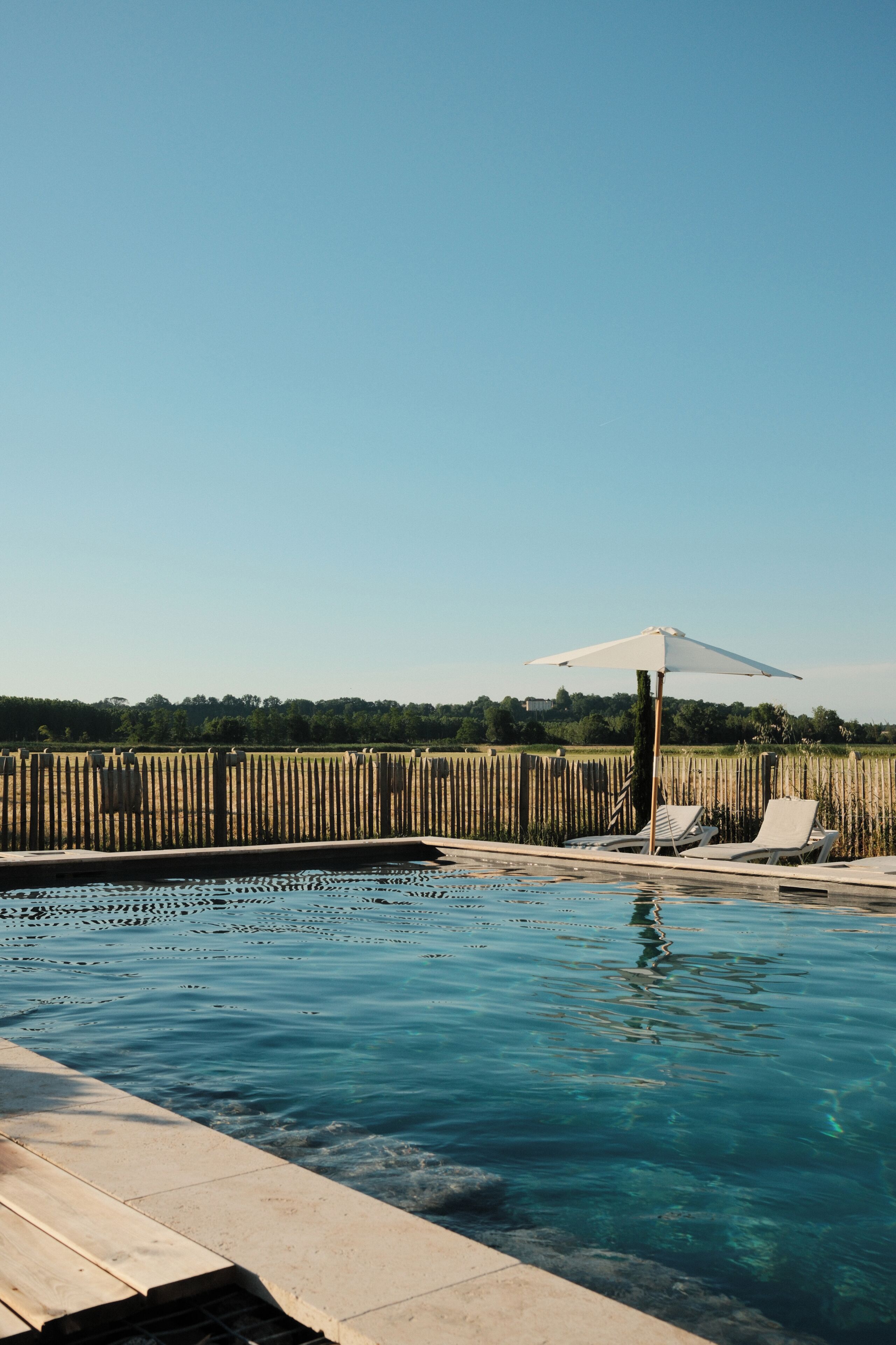 A clear blue swimming pool with two white lounge chairs and a white umbrella.