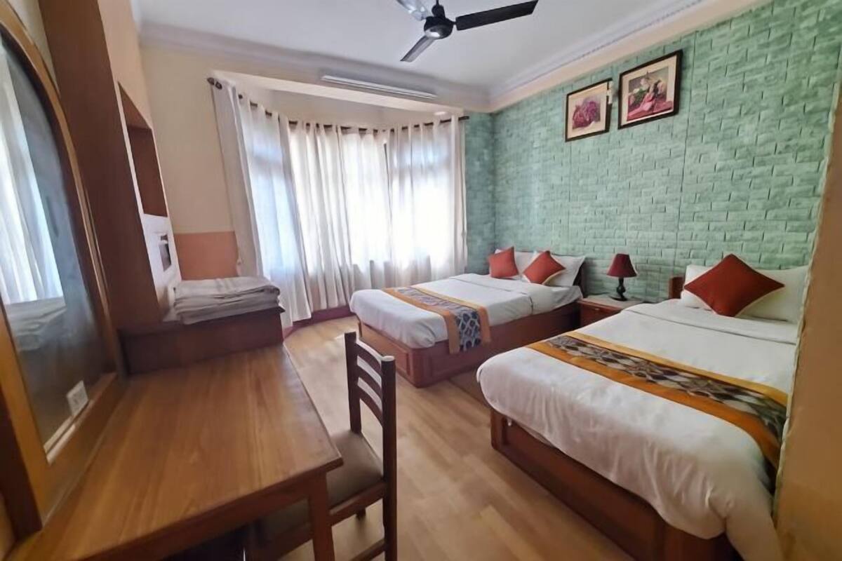 Una habitación de hotel con dos camas, un escritorio de madera, un ventilador de techo y una pared de ladrillo verde.