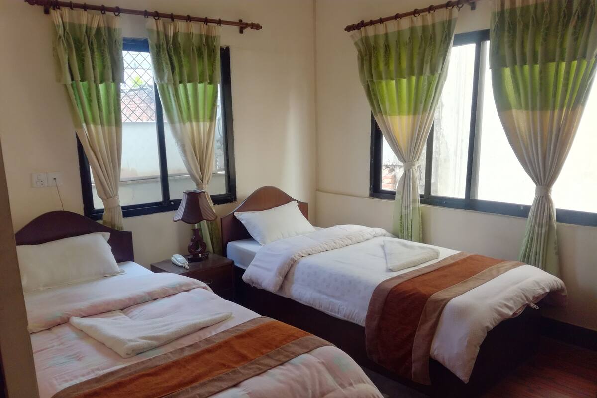 Habitación de hotel con dos camas individuales, una mesita de noche con lámpara y una ventana con cortinas verdes.