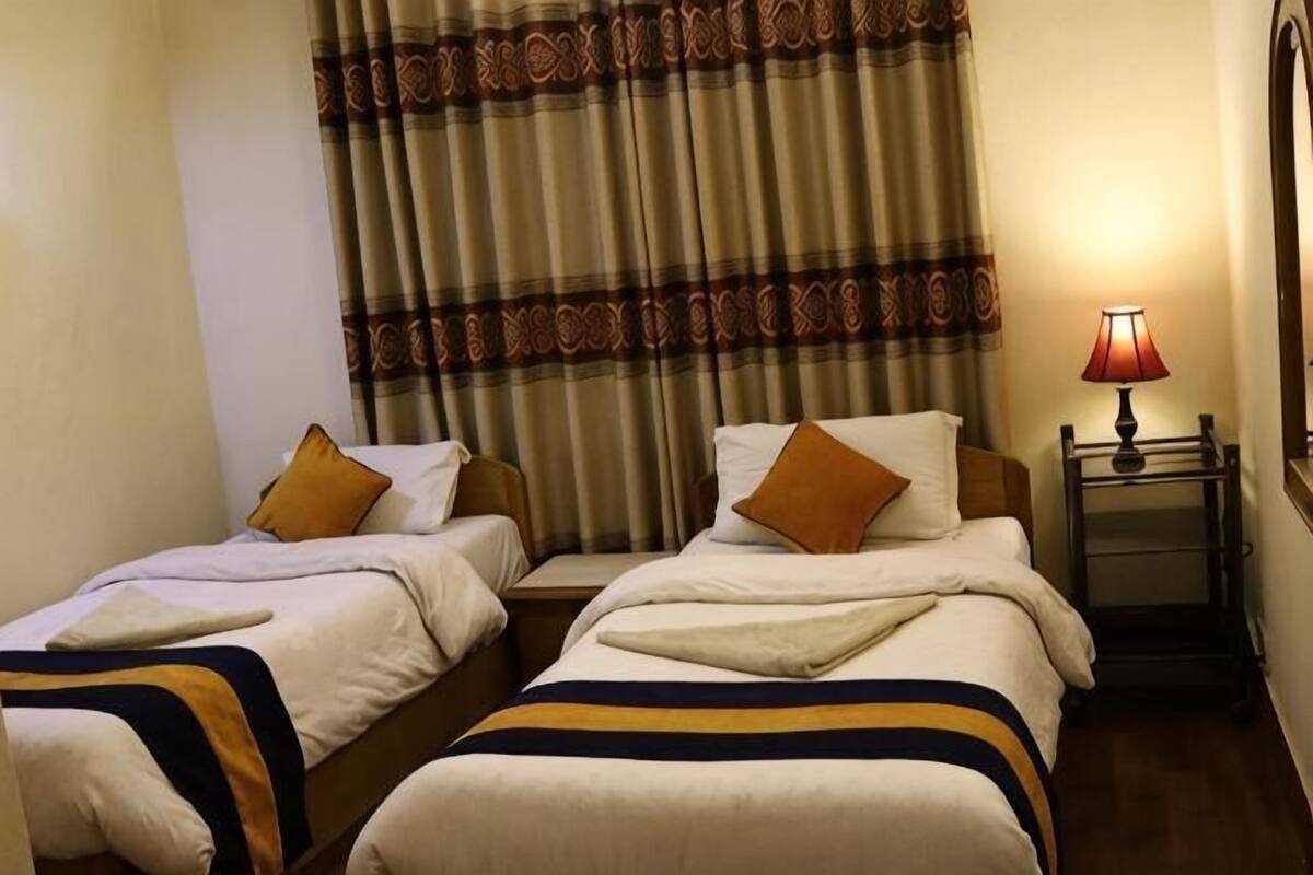 Habitación de hotel con dos camas, una mesita de noche con lámpara y una cortina estampada.