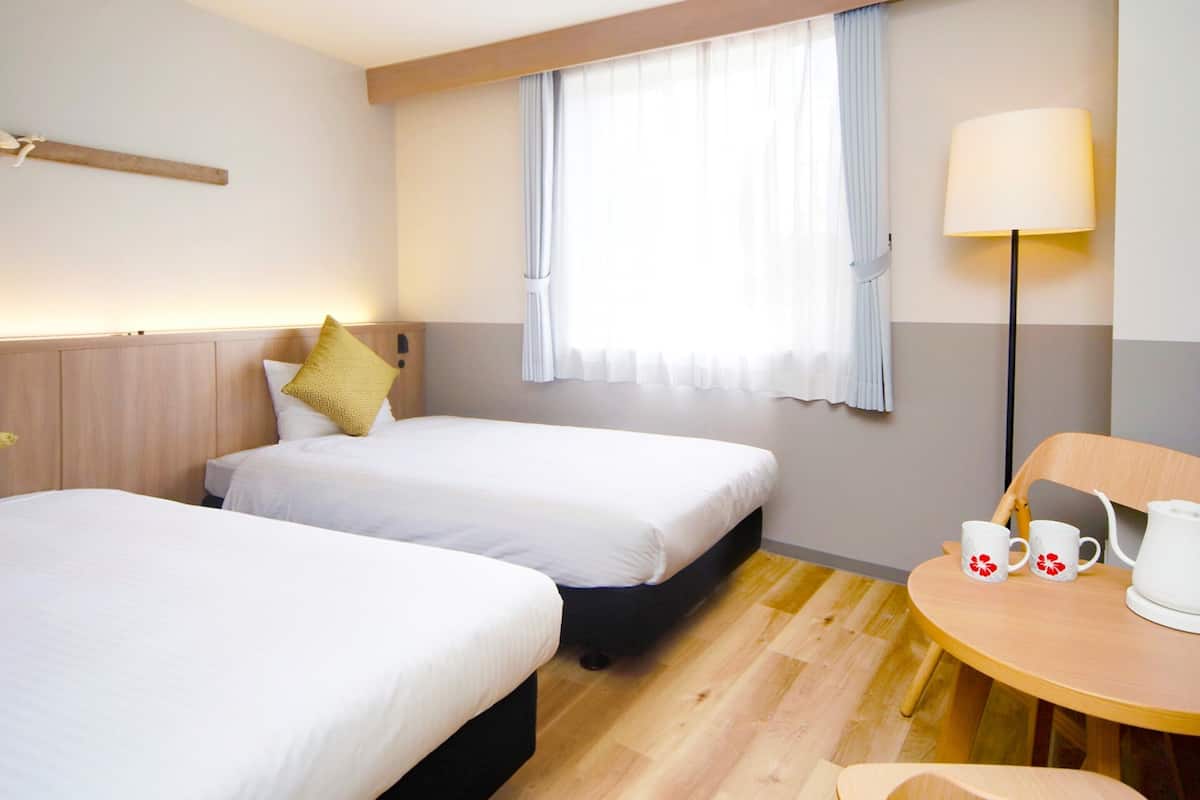 A hotel room with two beds, a small table with a lamp, and a window with curtains.