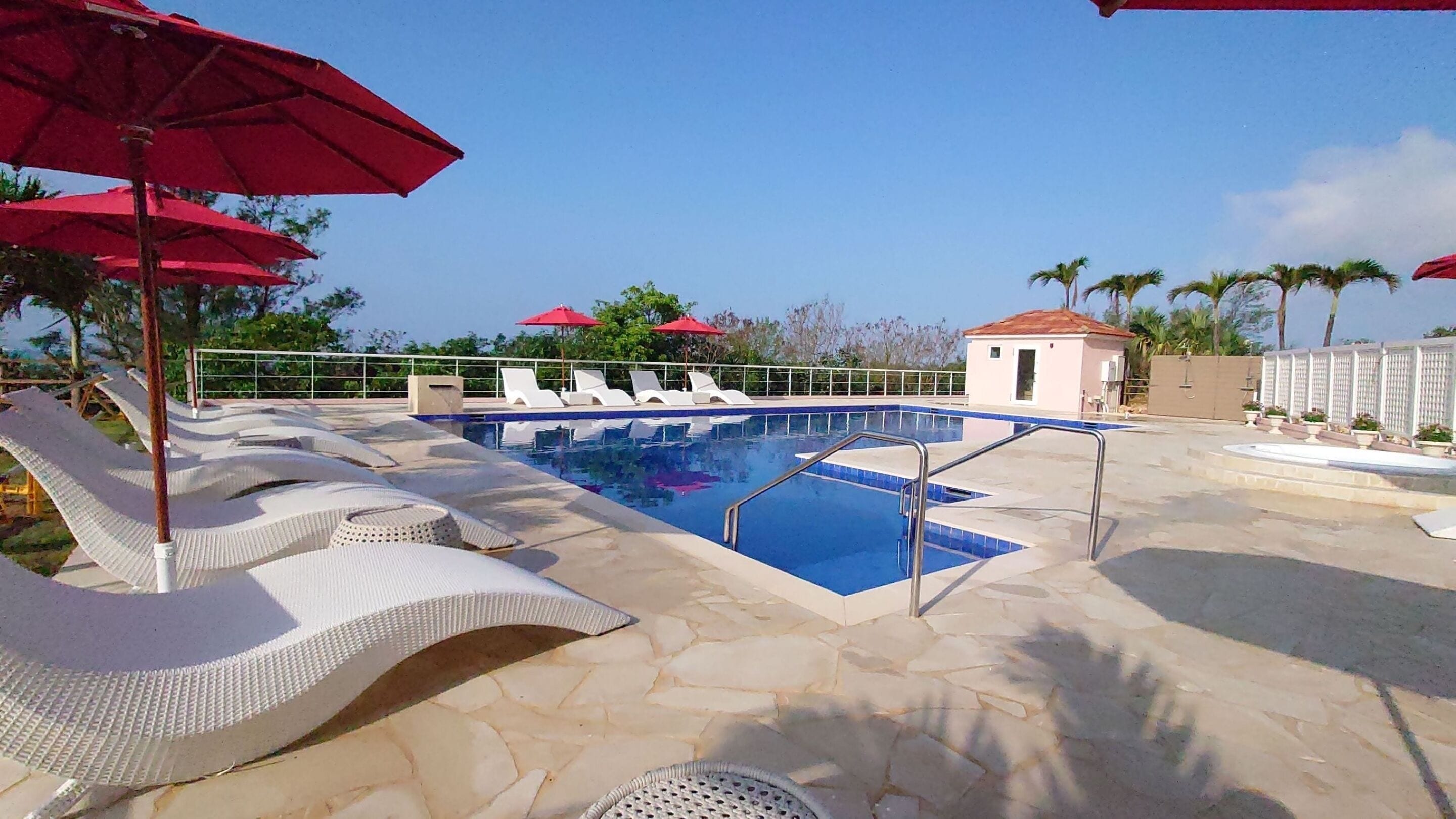 Une piscine entourée d’une terrasse en pierre et de palmiers, avec des chaises longues et des parasols rouges.