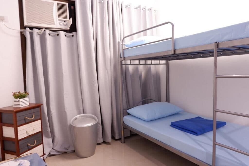 Un lit superposé avec un oreiller bleu, une petite commode en bois, une poubelle et un rideau.