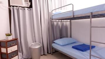 Un lit superposé avec un oreiller bleu, une petite commode en bois, une poubelle et un rideau.
