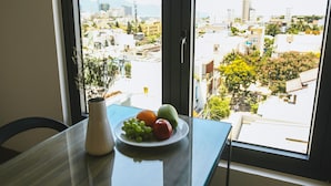 A dining table with a plate of fruit and a vase, a window with a view of a cityscape and mountains. - Red Hotel Da Nang (Da Nang)