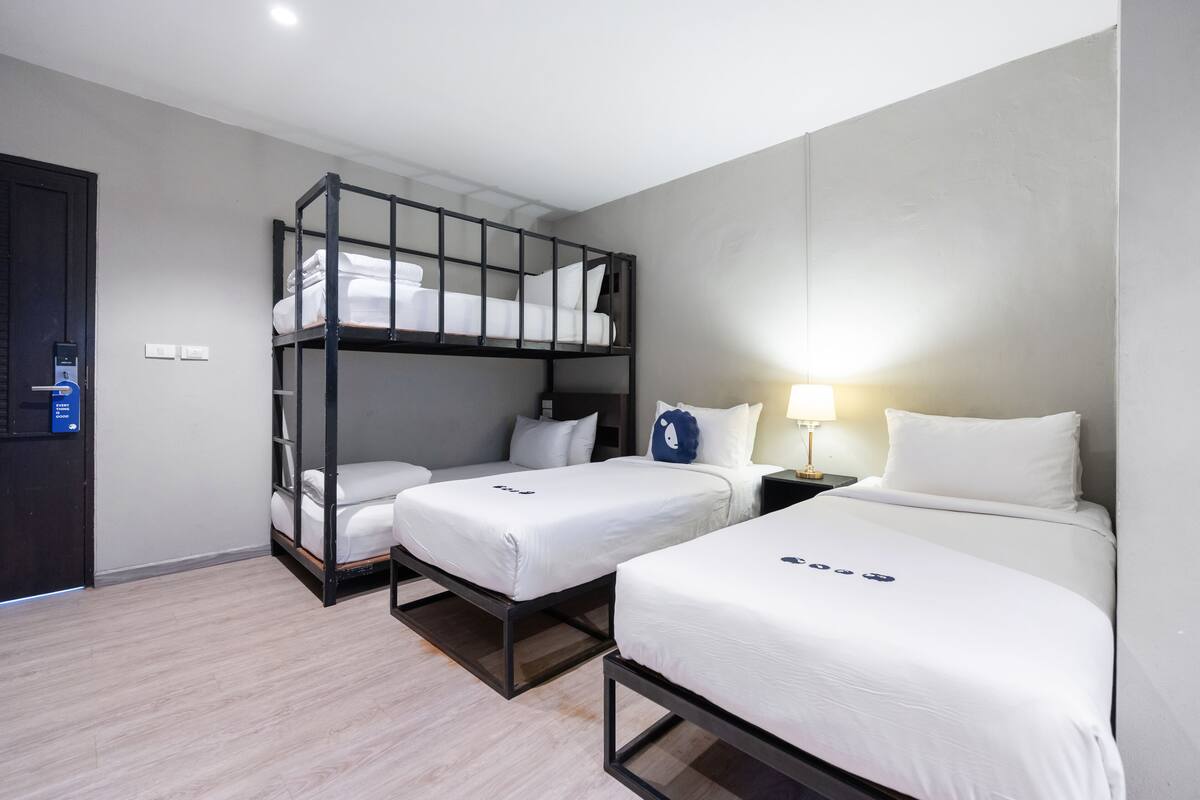 A hotel room with a bunk bed, two single beds, a bedside table with a lamp, and a door with a control panel.