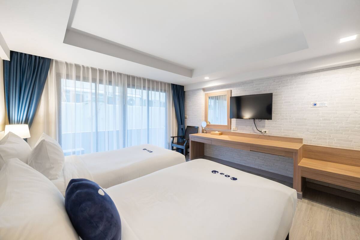 A hotel room with two beds, a desk, a TV, and a large window with curtains.