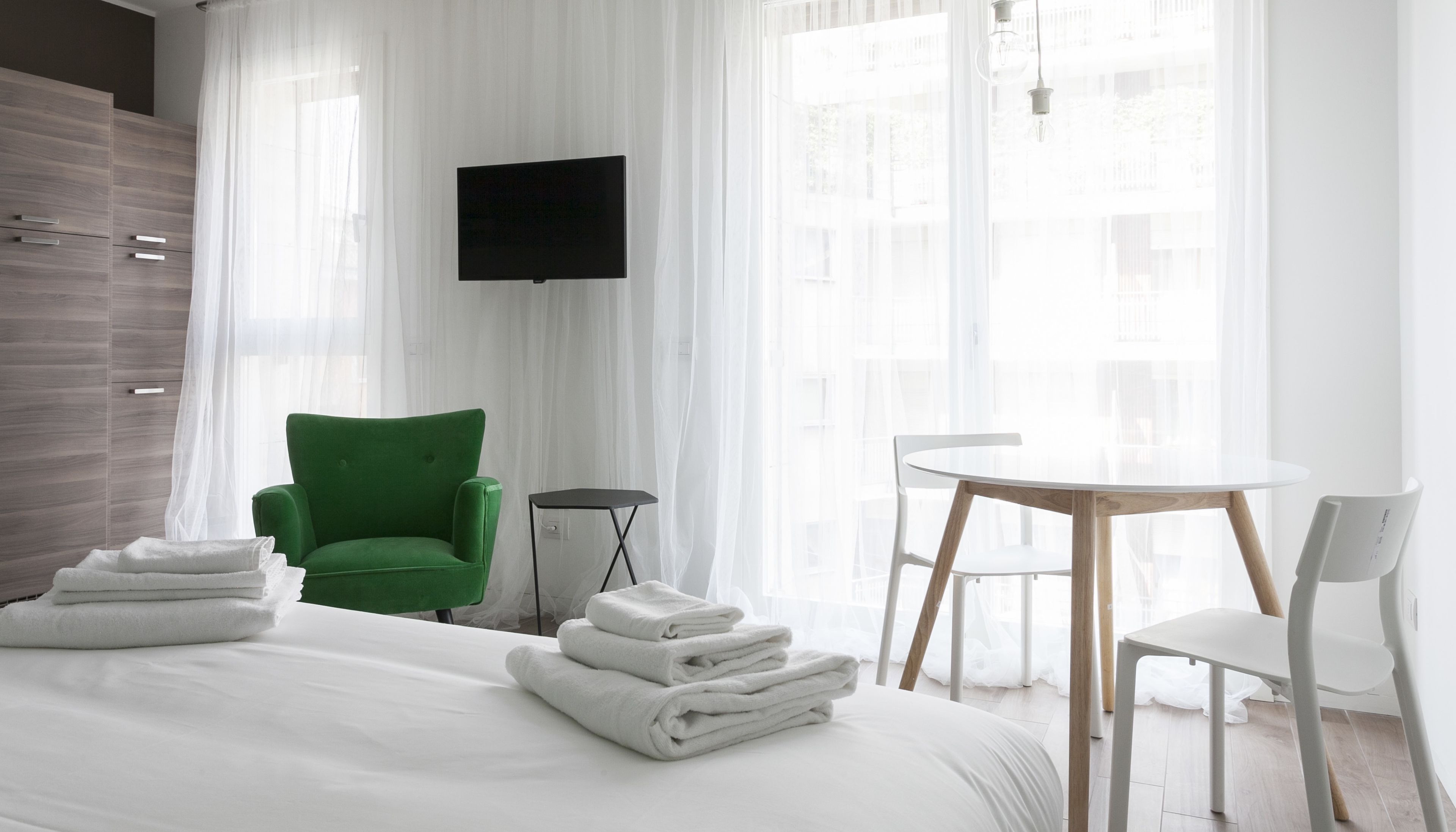 Una habitación de hotel con una cama, un sillón verde, una mesita pequeña, una silla blanca y un televisor montado en la pared.