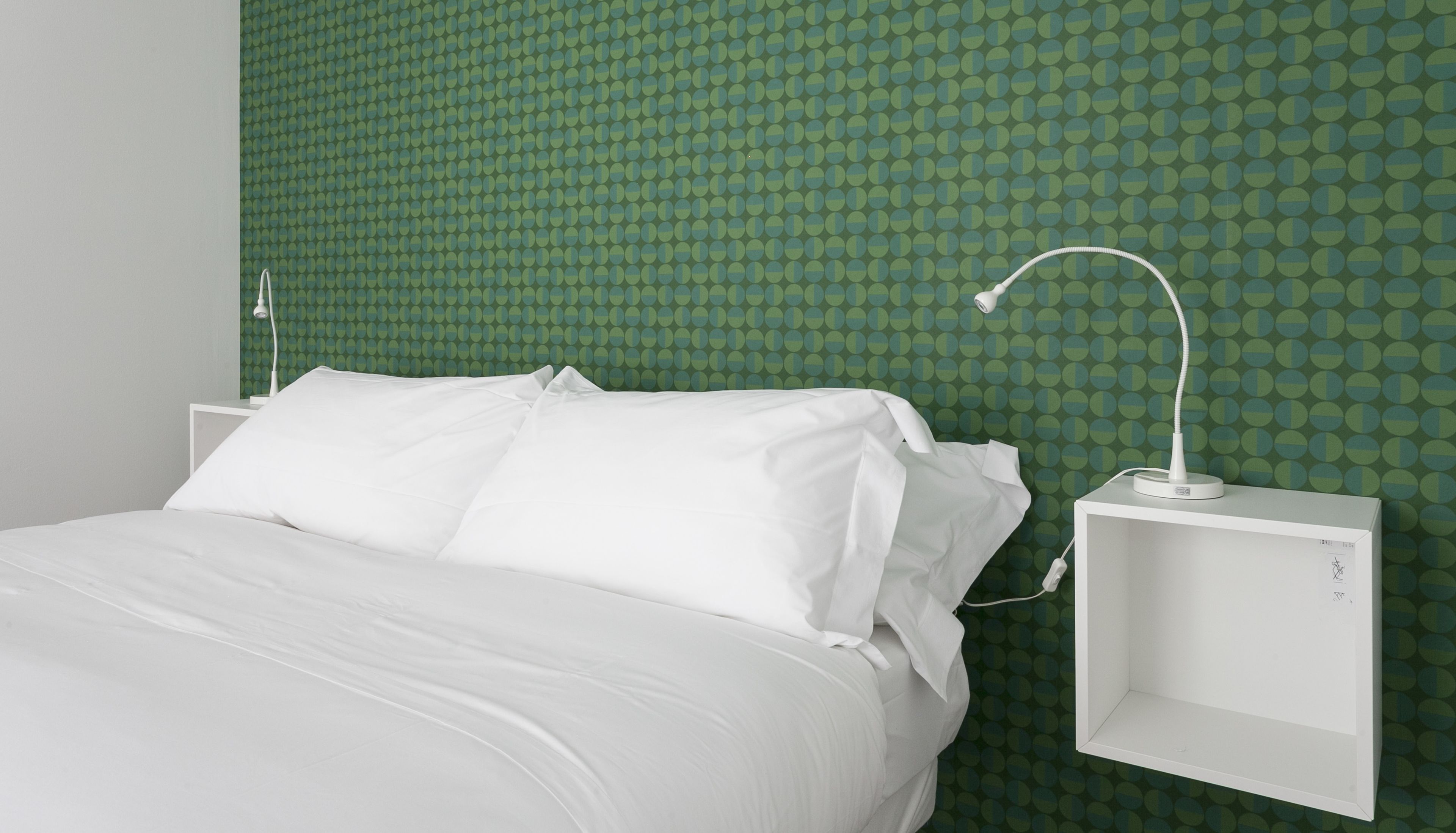 Een netjes opgemaakt bed met witte lakens in een kamer met behang met groene patronen en een witte nachtkastje met een lamp.