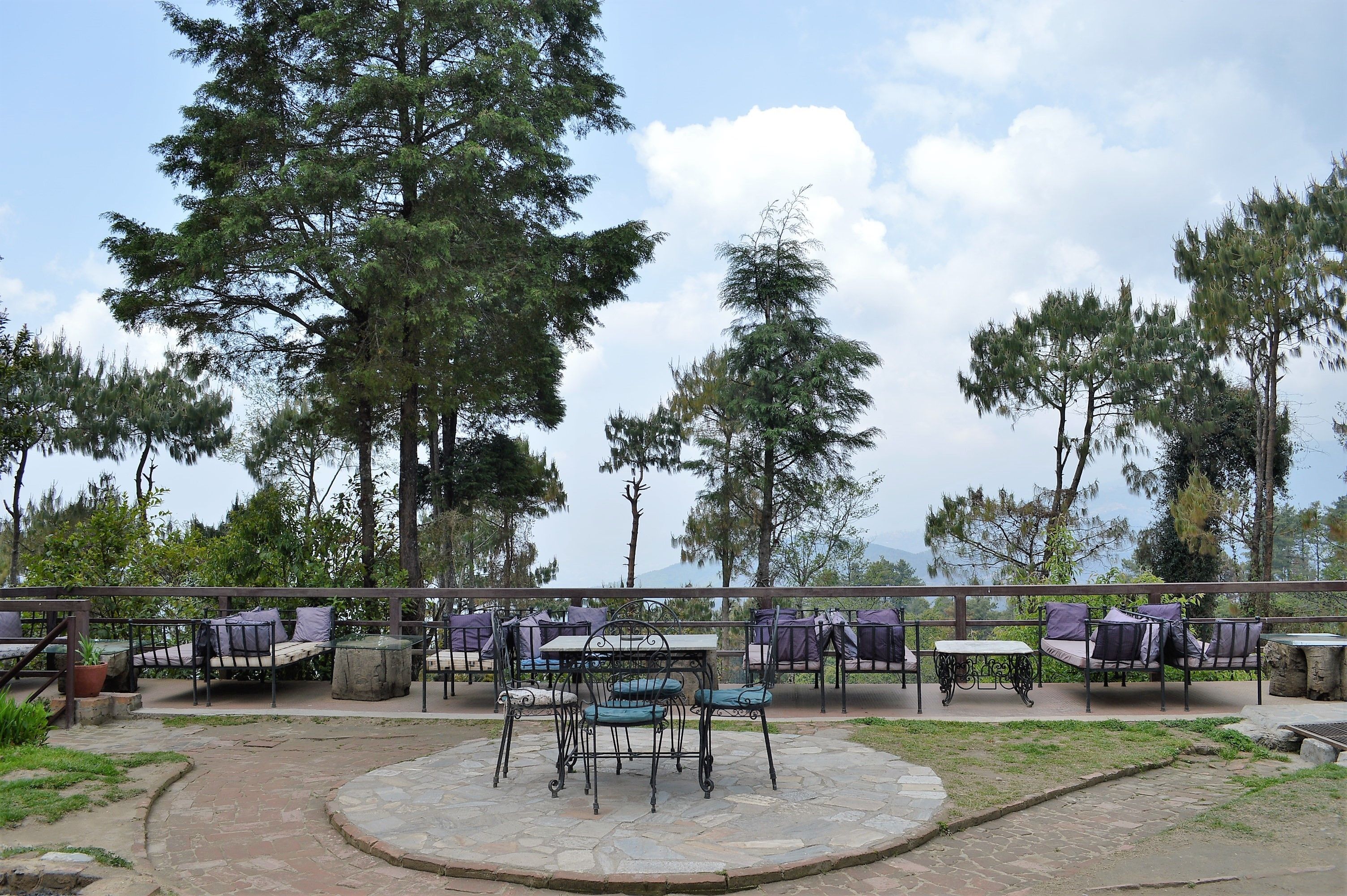 Un patio al aire libre con arreglos de asientos y una mesa central, rodeado de árboles y un sendero de piedra.