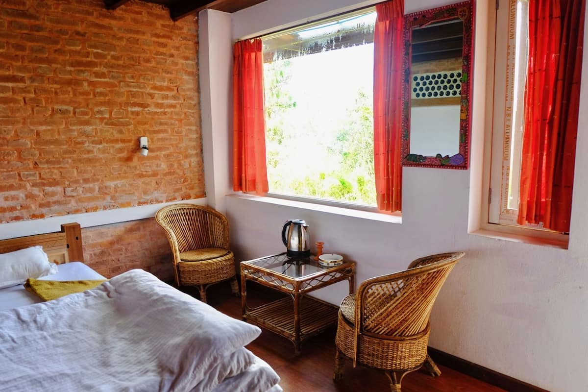 A room with a bed, two wicker chairs, a small table with a kettle, and a window with red curtains.