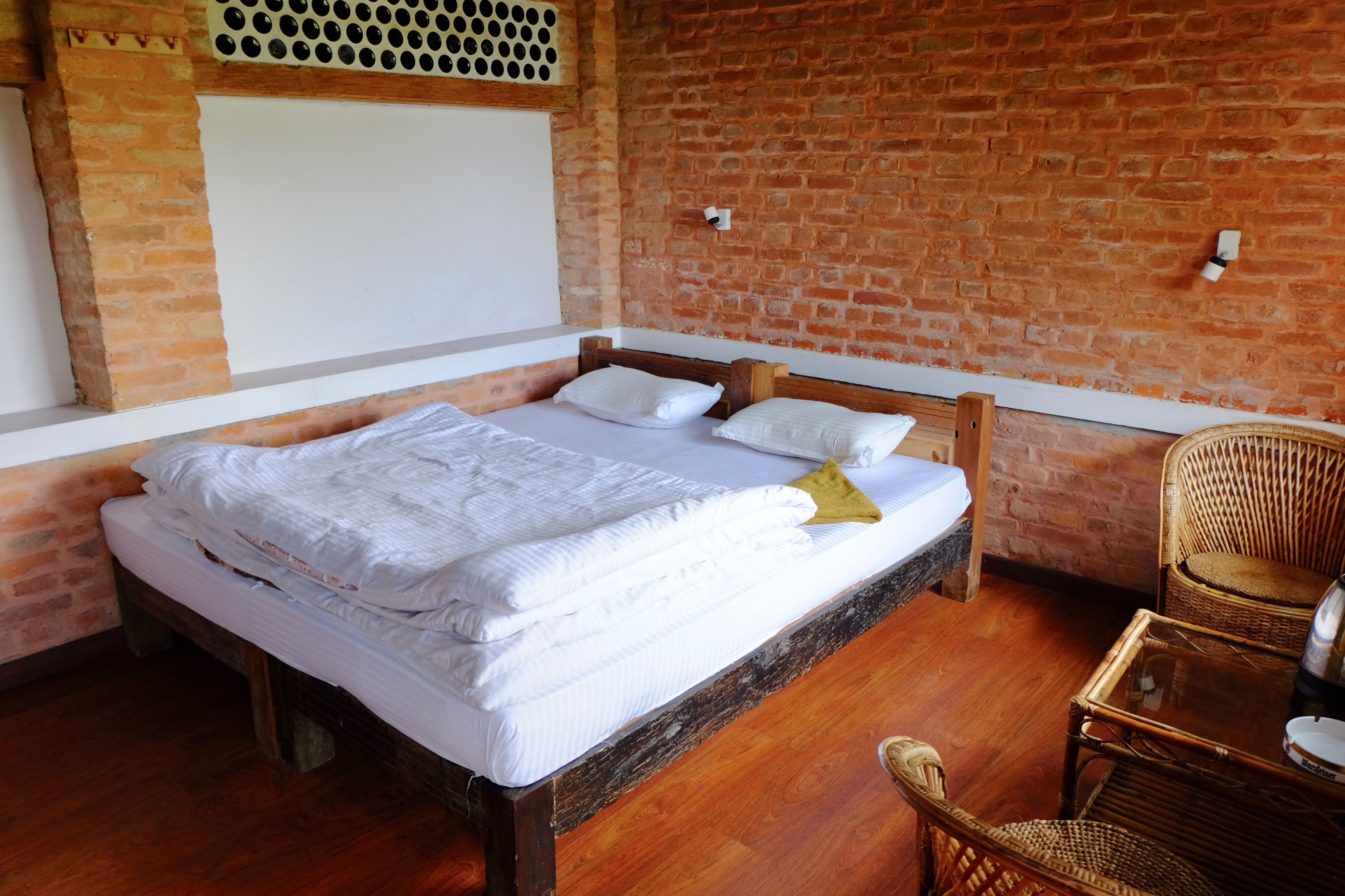 Una habitación con pared de ladrillo, cama de madera con ropa de cama blanca y una silla de mimbre.