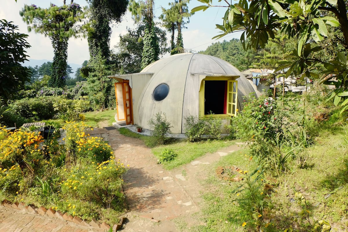 A dome-shaped building with a circular window and a door, surrounded by lush greenery and a garden path.