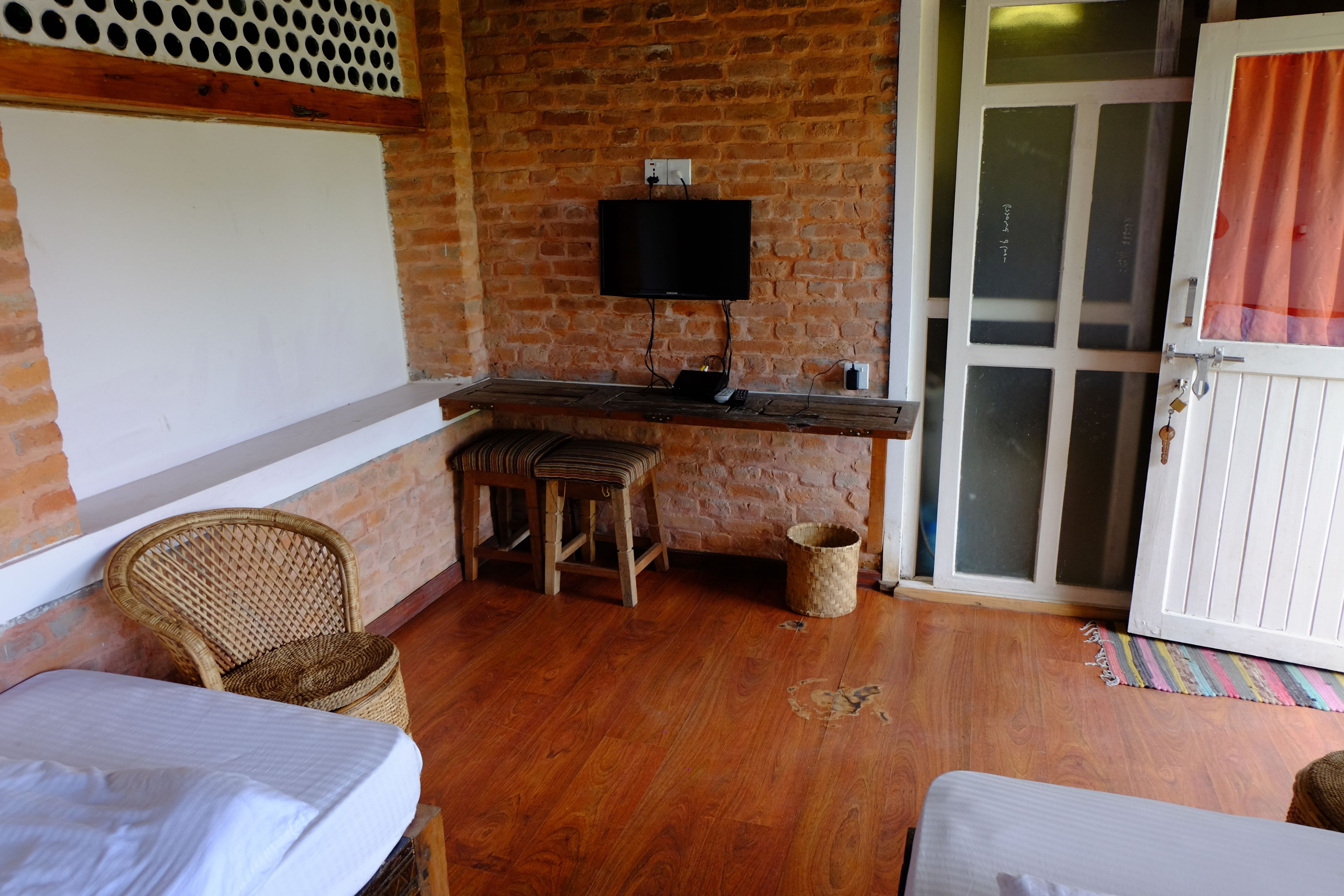A room with a brick wall, a flat-screen TV mounted above a wooden table, two wicker chairs, a small trash bin, and a door with a glass panel.