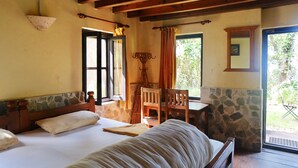 Una stanza accogliente con un letto, un tavolo di legno e una sedia. Si nota una finestra con tende, un muro in pietra e una vista sul verde circostante.