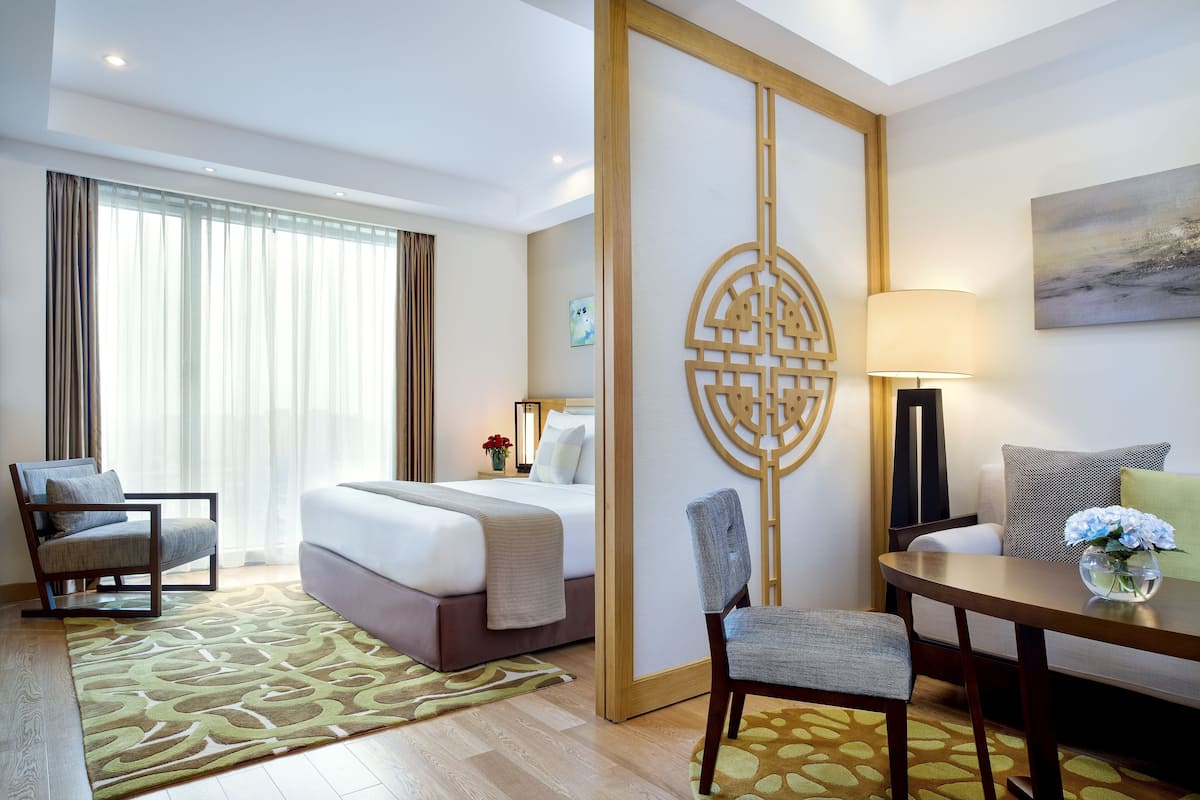 A hotel room with a bed, a desk, a chair, a sofa, a decorative screen, and a painting on the wall.