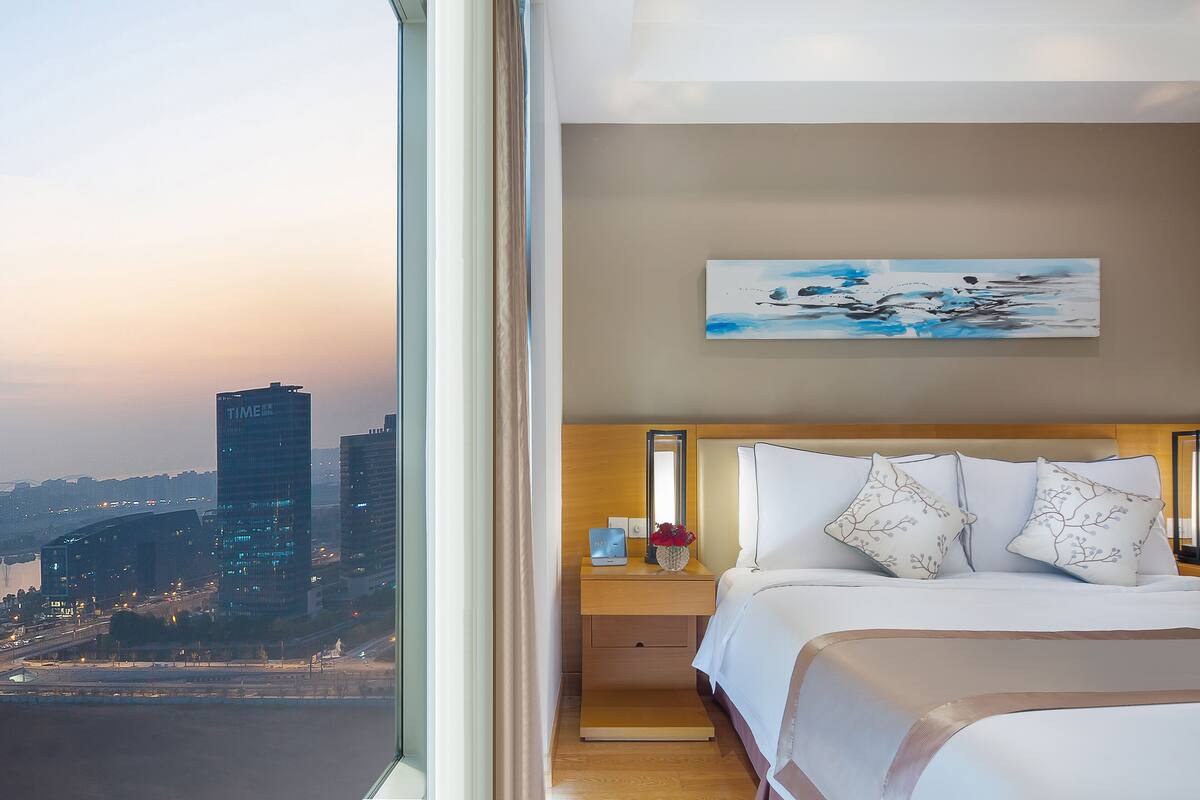 A hotel room with a large window, a bed with white bedding, a nightstand with a vase of flowers, and a painting on the wall.