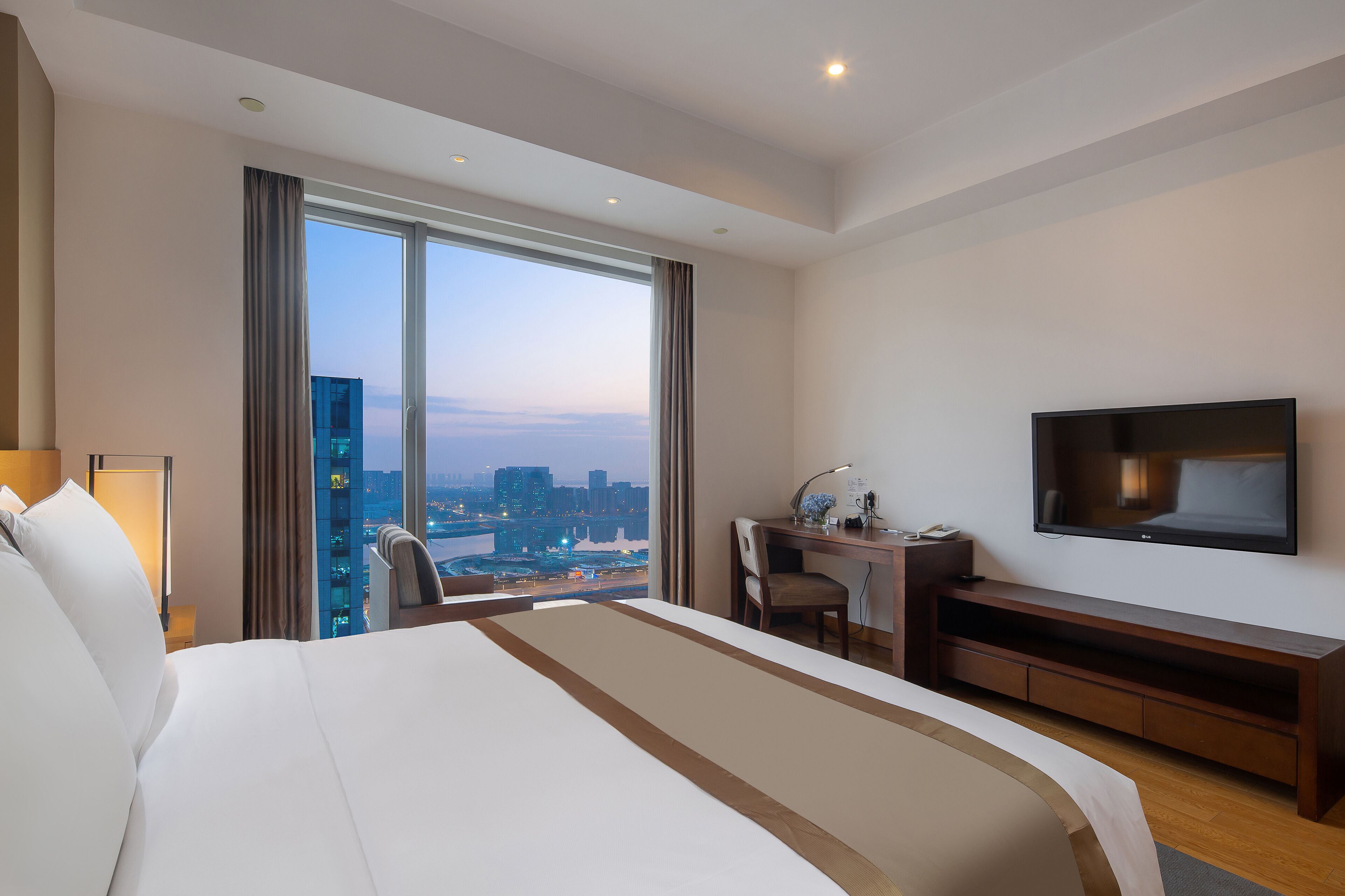Habitación de hotel con una cama grande, un escritorio con silla, un televisor de pantalla plana y vistas a un paisaje urbano a través de ventanas amplias.