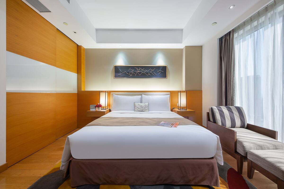 A modern hotel room with a large bed, bedside tables, a reading lamp, and a view of the city through the window.