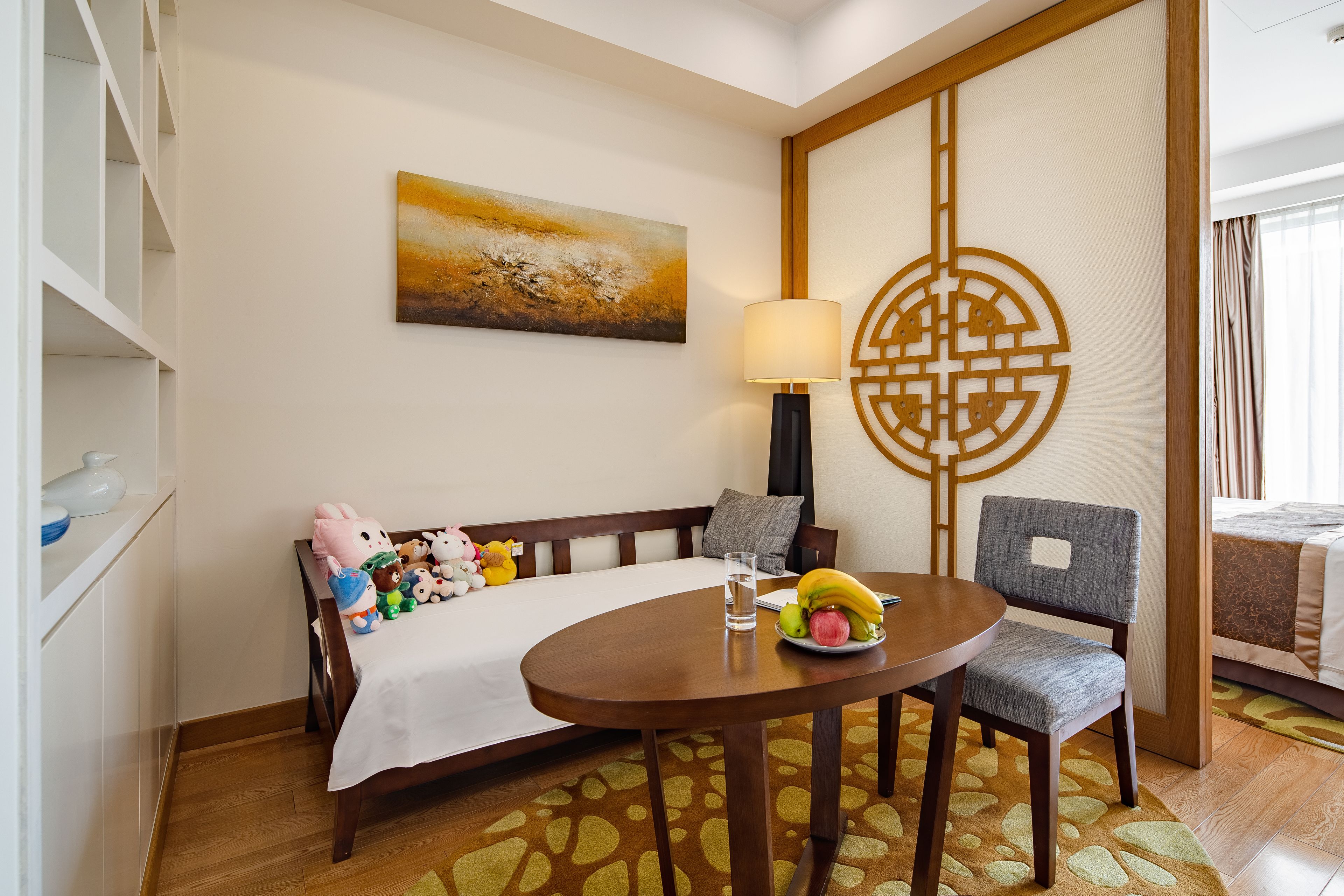 A hotel room with a bed, a dining table with a fruit plate, a lamp, and a decorative wall piece.