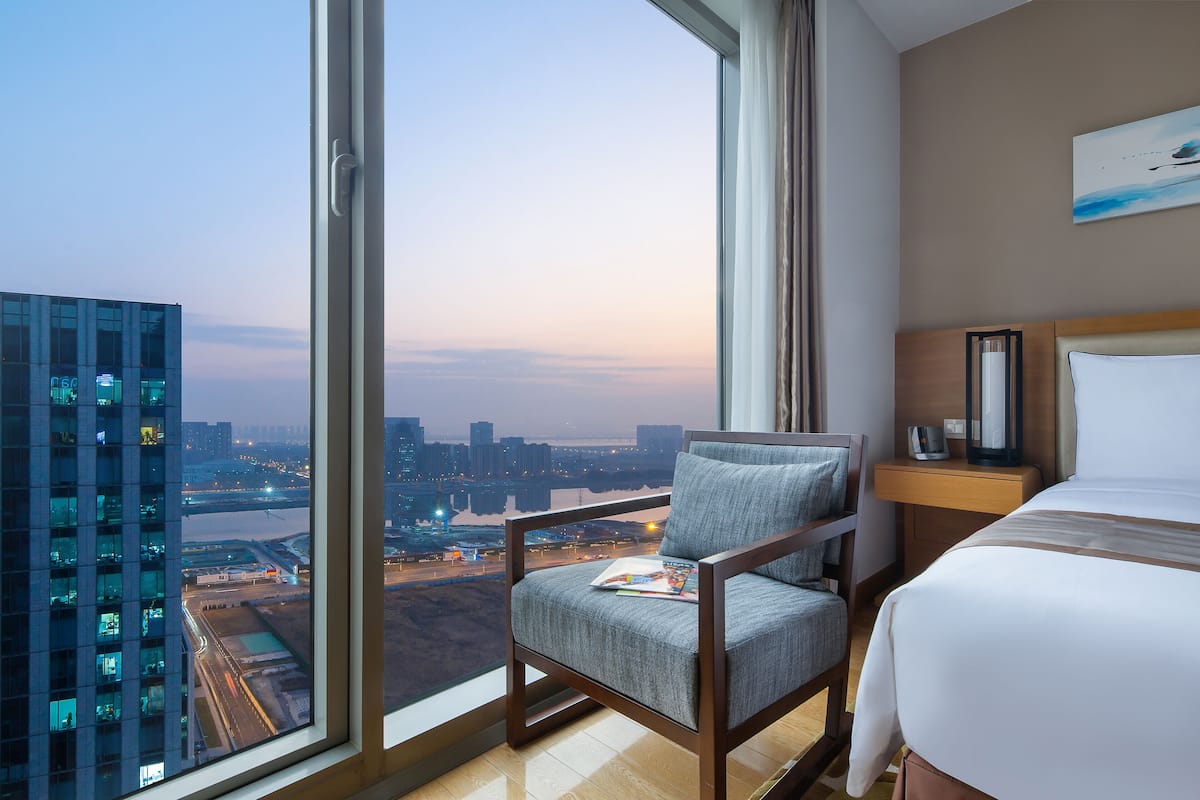 A hotel room with a large window offering a city view, a bed, a chair, and a nightstand.