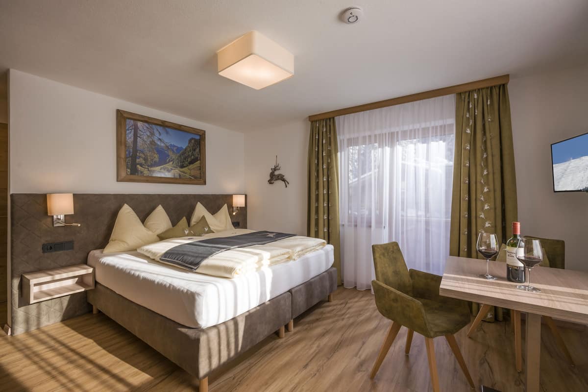 Una habitación de hotel con cama, mesita de noche, lámpara, cortinas, un cuadro, una cabeza de ciervo montada y un televisor en la pared.