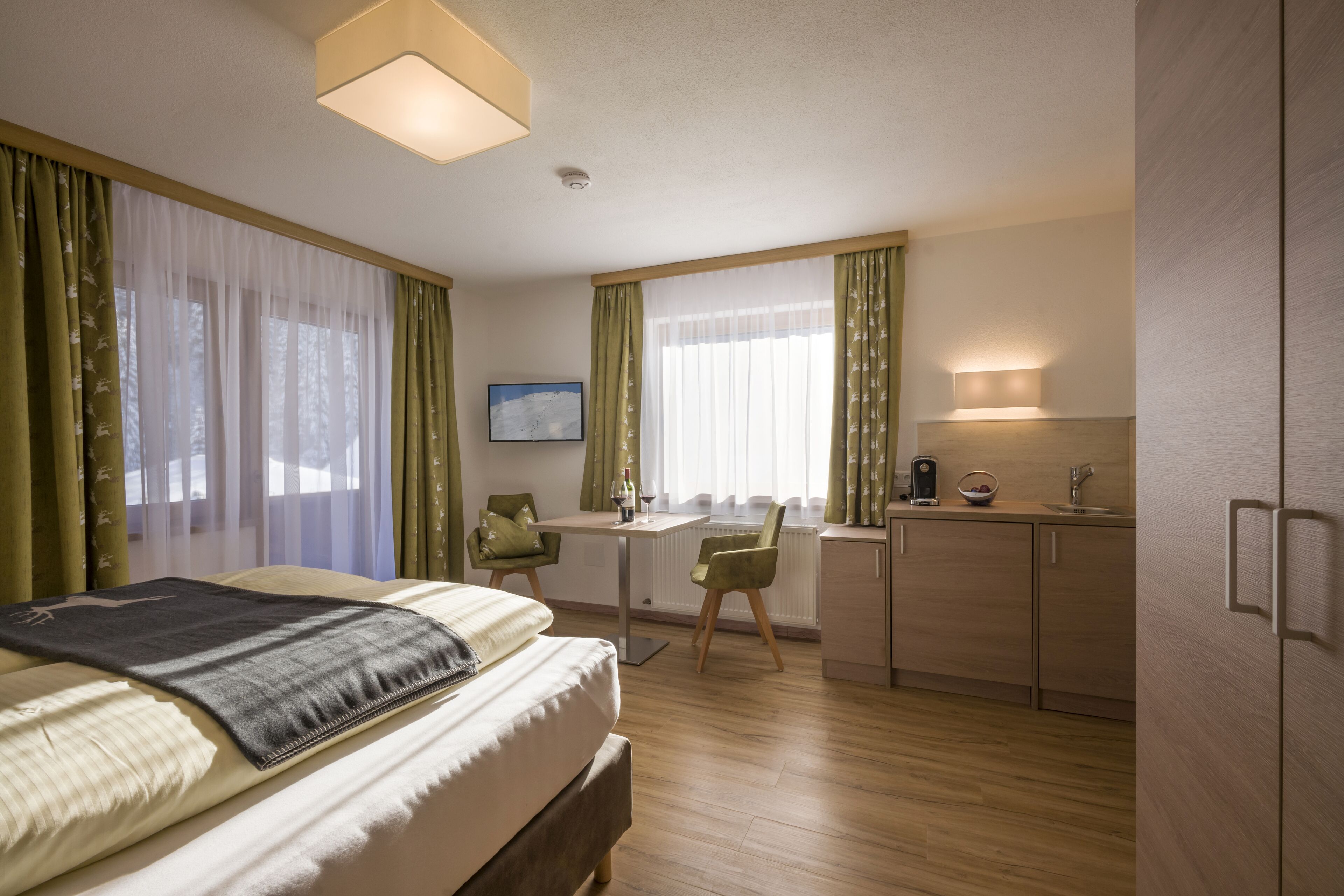 A hotel room with a bed, a desk, a chair, a small kitchenette, and a window with curtains.
