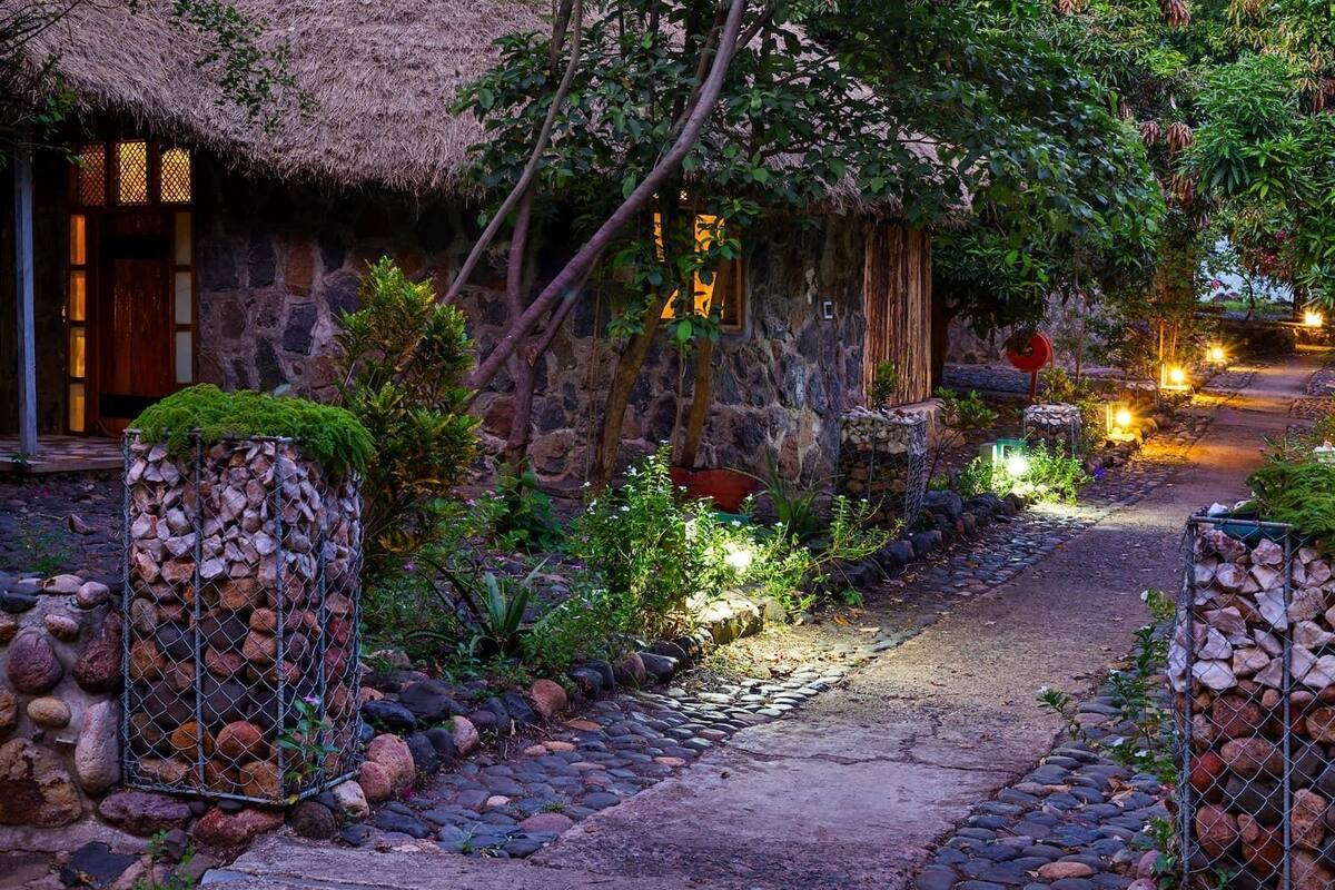A stone pathway leading to a rustic building with a thatched roof, surrounded by lush greenery and illuminated by warm lights.