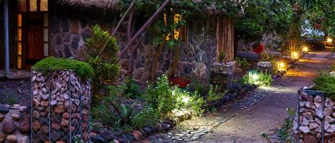 A stone pathway leading to a rustic building with a thatched roof, surrounded by lush greenery and illuminated by warm lights.