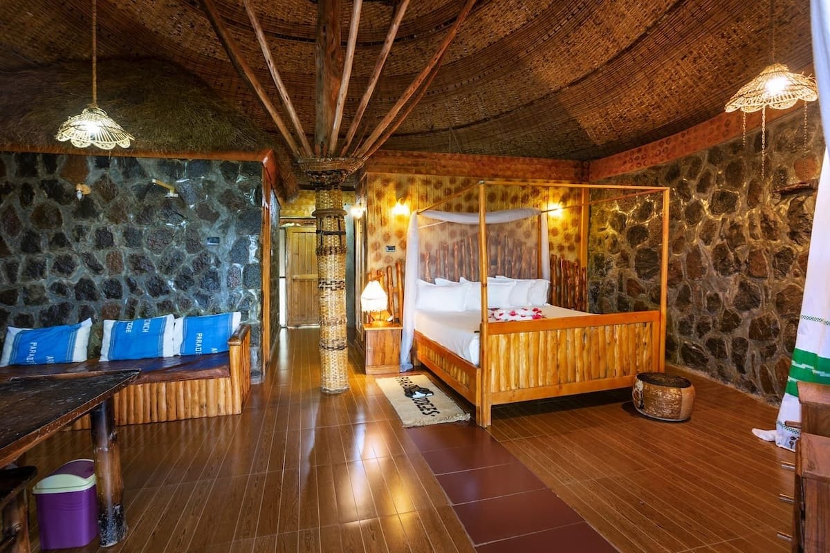 A rustic room with a stone wall, a wooden four-poster bed, a bench with cushions, and a wooden floor.