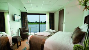 A hotel room with a bed, a chair, a small table, and a view of a body of water through a large window. - Crossgates Hotelship AltstadtNord - Köln (Cologne)