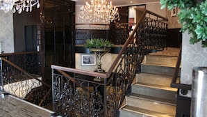 A grand staircase with ornate metal railings and a chandelier, leading to a landing with a potted plant and framed picture. - Crossgates Hotelship AltstadtNord - Köln (Cologne)