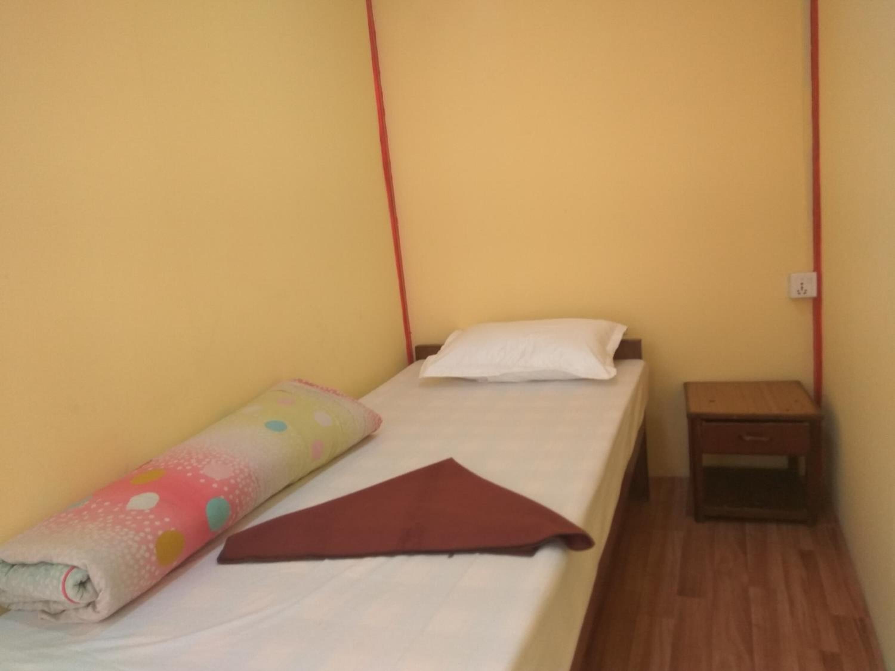 A small, single-bed room with a wooden floor, a bedside table, and a wall socket.