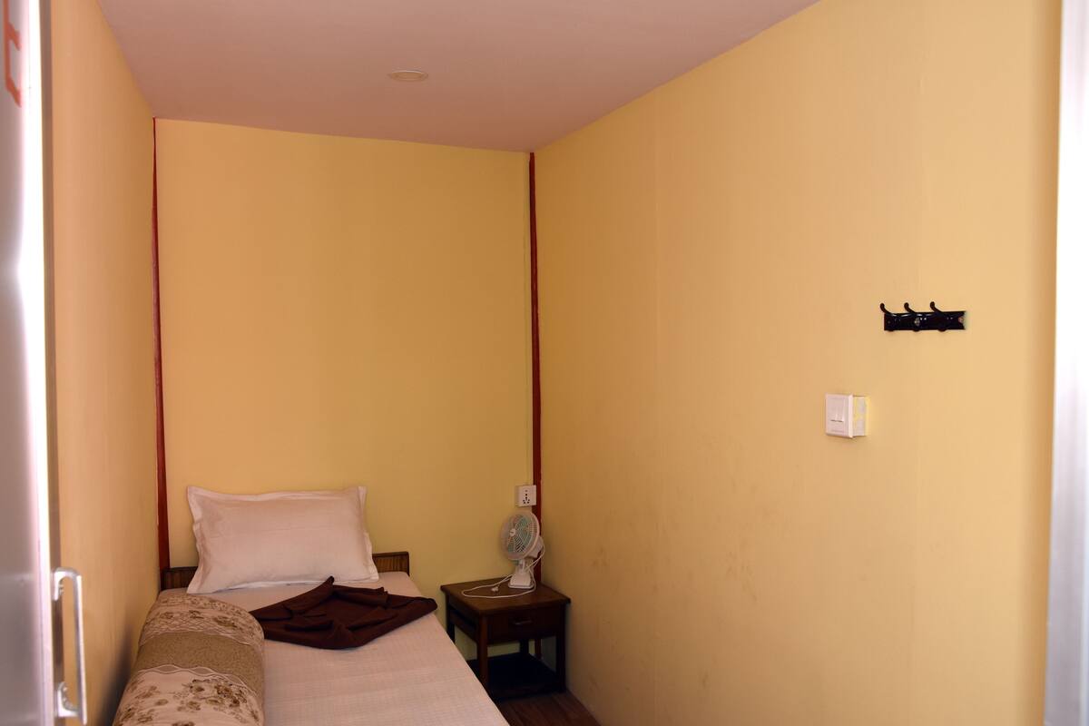 A small, single-bed room with a wooden headboard, a bedside table with a fan, and a wall-mounted light switch.
