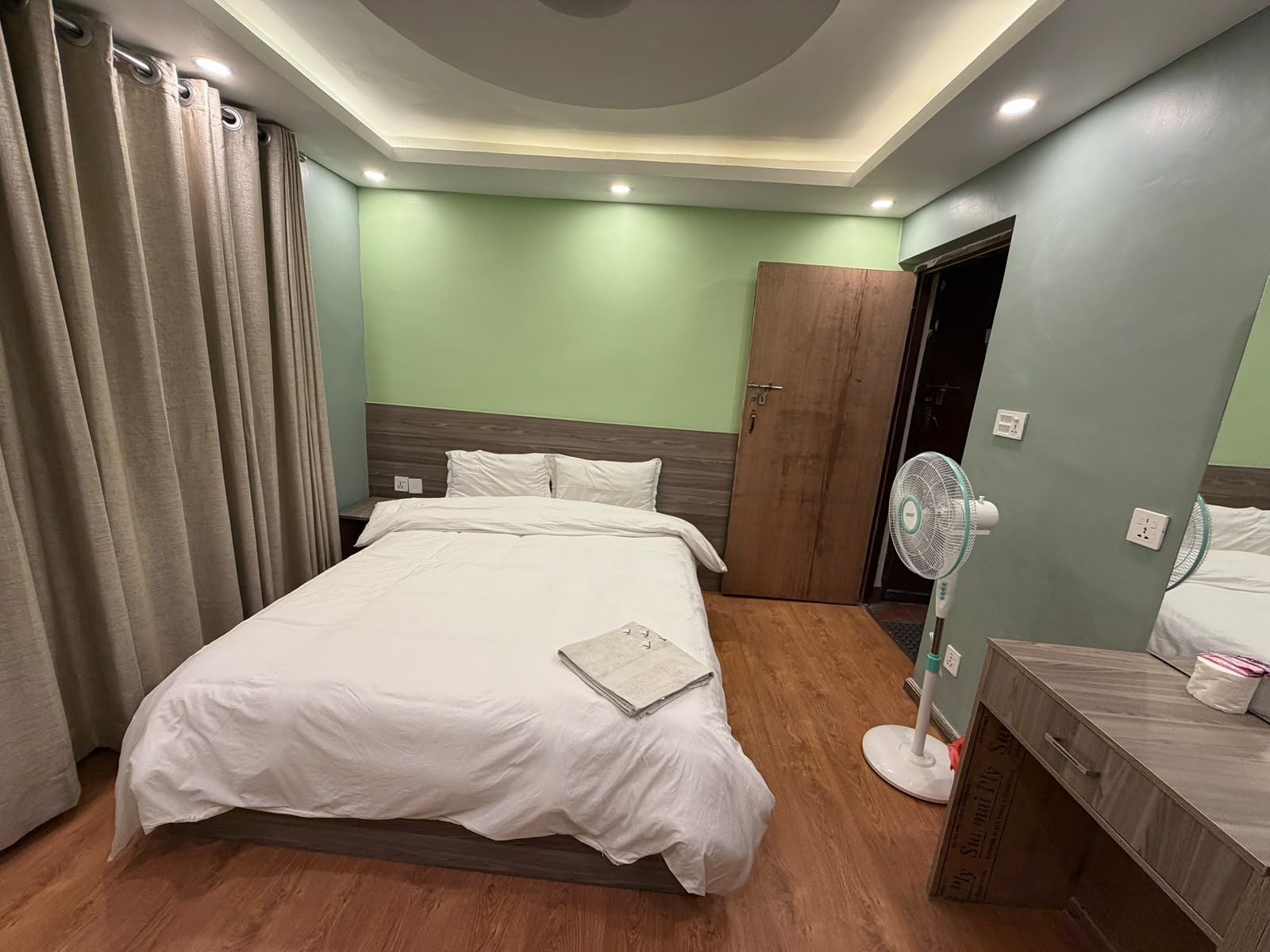 Une chambre d’hôtel avec un lit, un ventilateur de chevet, une armoire en bois et un miroir.