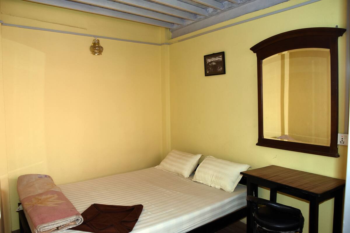 A small, single-bed room with a wooden nightstand, a mirror, and a wall-mounted light fixture.