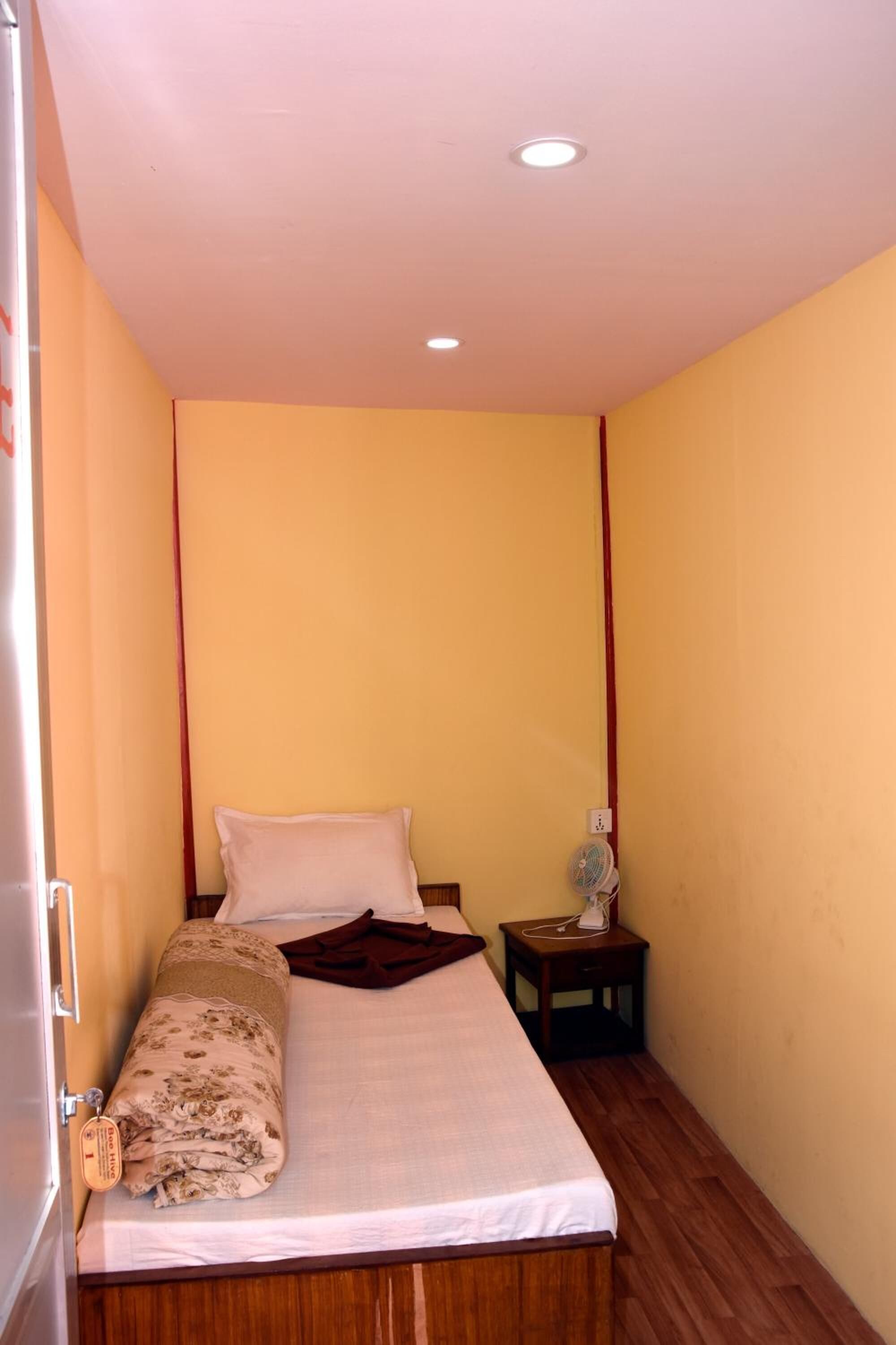 A small, single-bed room with a wooden floor, a bedside table with a lamp, and a wall-mounted fan.