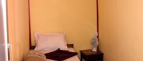 A small, single-bed room with a wooden floor, a bedside table with a lamp, and a wall-mounted fan.