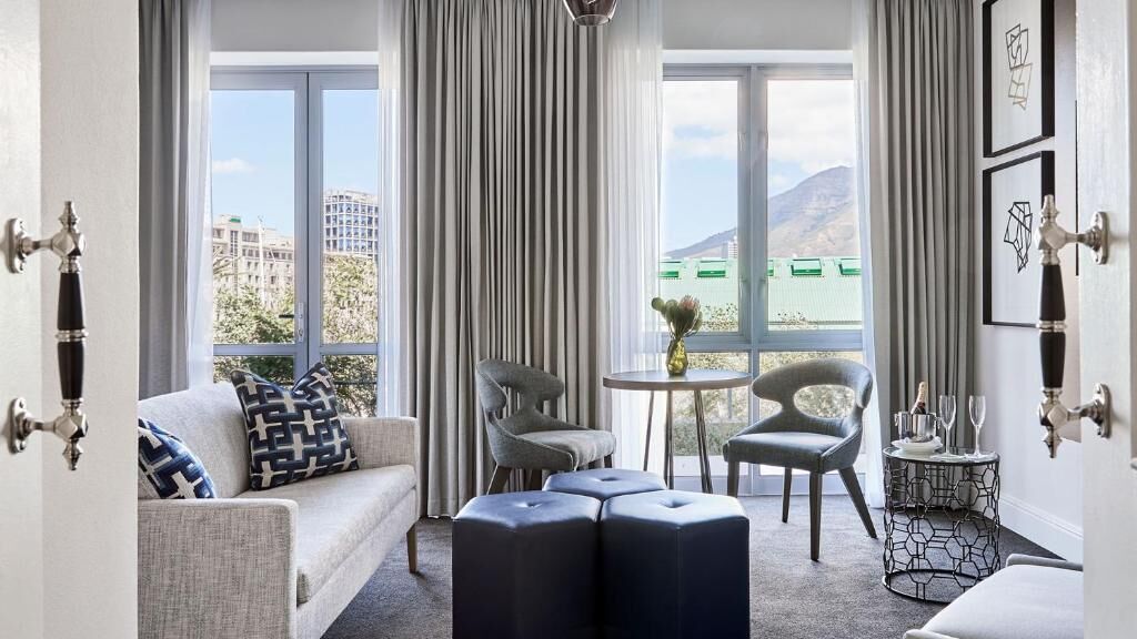 A hotel room with a sofa, two chairs, a small table with a vase, and a view of buildings and mountains.