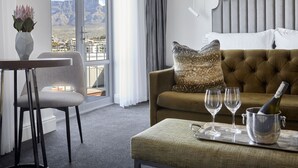 A modern hotel room with a view of a mountain range, a sofa with a textured pillow, a dining area with a chair, and a table with a bottle of champagne and glasses.